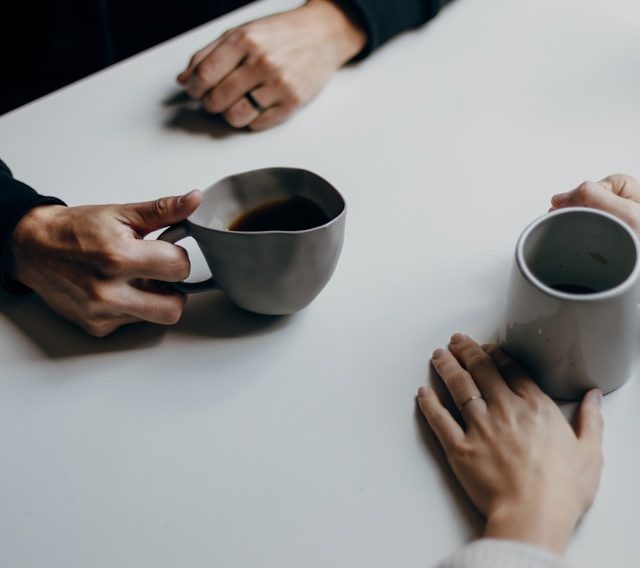 Two people sit across from each other at a table, each holding a mug, with only their hands visible. The scene is minimal with neutral tones.